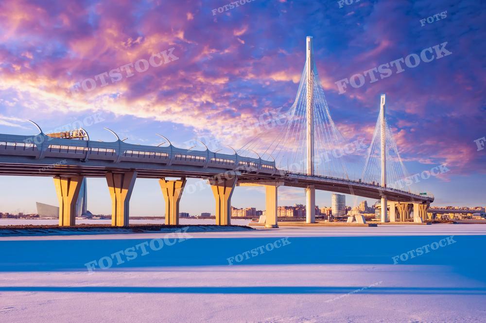 На фотографии запечатлен Вантовый мост через реку Нева в Санкт-Петербурге на фоне впечатляющего заката. Небо окрашено в теплые оттенки оранжевого и розового, что контрастирует с холодными синими тонами снега на переднем плане. Фото может быть использовано для иллюстрации статей о современной архитектуре, в туристических публикациях или как вдохновение для художников.
